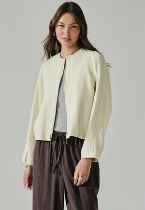 Young woman with long dark hair wears cream zip-up jacket over gray top and loose dark brown pants, standing against neutral background.