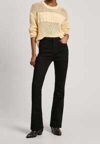 Vaqueros negros acampanados combinados con un suéter de punto amarillo dorado con un patrón texturizado y un ajuste holgado. Botines hasta el tobillo completan el conjunto.