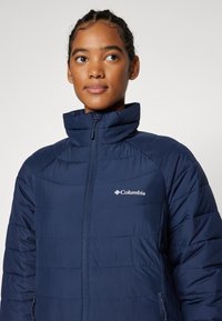 Giacca imbottita blu navy con colletto alto, design trapuntato e zip completa. Presenta un logo Columbia sul petto sinistro e tasche laterali.