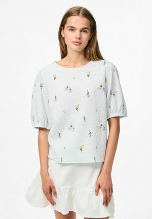 Ljusblå blommönstrad topp med korta puffärmar, med en texturerad tyg och rund halsringning, ihop med en vit rutig kjol.