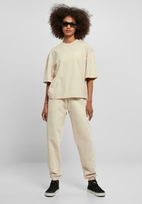 Beige oversized tričko a odpovídající jogger kalhoty z měkkého materiálu. Tričko má krátké rukávy, jogger kalhoty disponují elastickým pasem a manžetami. Černé sluneční brýle.