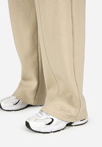 Krémszínű, texturált, széles lábú nadrág, amely waffle mintás, fehér edzőcipőkkel párosítva, fekete részletekkel és fűzőkkel.