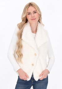 Blonde vrouw in een witte fuzzy mouwloze vest met gouden knopen over een witte lange mouwen top en blauwe jeans, met een zachte glimlach.