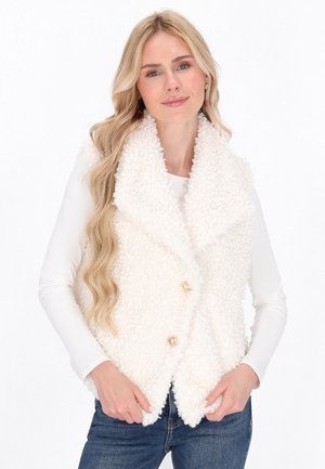 Blonde Frau, die eine weiße, flauschige ärmellose Weste mit goldenen Knöpfen über einem langärmeligen weißen Top und blauen Jeans trägt, und sanft lächelt.