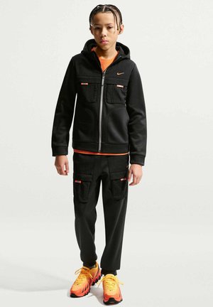 Giovane che indossa una felpa nera con zip, pantaloni neri con tasche, una maglietta arancione sotto e scarpe da ginnastica arancioni e gialle brillanti.