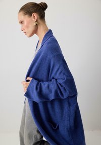 Naine, kellel on juuksed bunis, kannab kuldseid kõrvarõngaid ja ülekaalulist sinist kampsunit halli riietuse peal, seistes külg ees ühevärvilise tausta taustal.