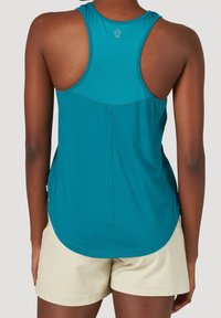 Kvinna som bär en teal racerback tanktop och krämfärgade shorts, sedd från ryggen mot en neutral bakgrund.