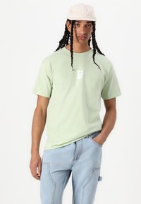 Lichtgroene katoenen t-shirt met een wit grafisch logo op de borst, gecombineerd met lichtblauwe denim broek met zijzakken.