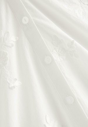 Hvidt stof med subtil blomsterbroderi og en lodret række af små hvide knapper til lukning.