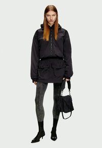 Casaco impermeável preto com fecho frontal, cintura ajustada e grandes bolsos, combinado com leggings cinzentas com padrão e botas de tornozelo pretas.