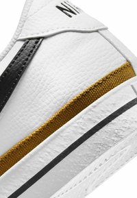 Sneaker bianco con una tomaia sintetica testurizzata, dettagli neri e dorati, e una suola in gomma con una decorazione a motivo.