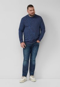 Marineblaues Sweatshirt mit Baumzweig-Muster, gerippten Bündchen und Rundhalsausschnitt, kombiniert mit ausgewaschenen blauen Jeans und weißen Turnschuhen.