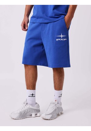 Man draagt blauwe PXP-shorts, witte sokken met PXP-logo en witte Nike-sneakers, staand met één hand in de zak.