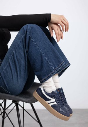 Zapatillas de marino con acentos blancos, suela de goma y puntera redondeada. El modelo lleva puestos unos vaqueros de pierna ancha azules y calcetines blancos de canalé.