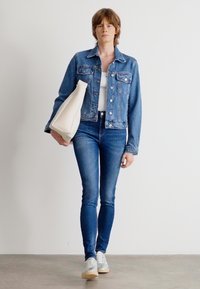 Farmerdzseki elülső zsebekkel, fehér ujjatlan felső, kék skinny farmernadrág és szürke sportcipő. A modell egy fehér, sima textúrájú táskát tart.
