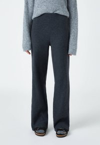 Pantalon large taille haute gris foncé en tissu doux et texturé, associé à un pull en maille gris ample et des sandales noires à enfiler.
