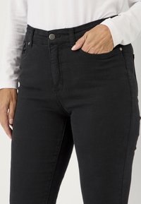 Mustat stretch-farkut, joissa on istuva muotoilu, etutasku, nappisuljenta ja koristeellinen ommel, käsi taskussa.