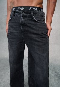 Pantalones de mezclilla negros con un corte relajado, que cuentan con una cinturilla negra de marca. Bolsillos frontales con detalle de costura visible. Sin patrón.