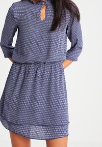 Dunkelblaues Kleid mit geometrischem Muster, Dreiviertelärmeln, Schlüsselloch-Ausschnitt und elastischem Taillengummizug mit leichtem Rüschensaum.