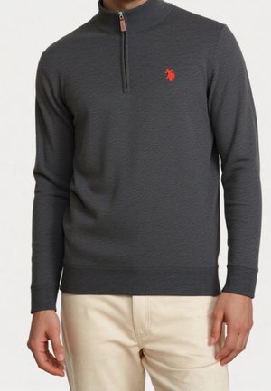 Mężczyzna noszący ciemnoszary pulower z długim rękawem i kołnierzem na zamek do ćwiartki, z małym czerwonym wyszywanym logo, w połączeniu z jasnobeżowymi spodniami.