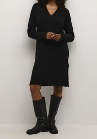 Robe en maille noire avec un col en V, des manches longues et des fentes latérales. Associée à des bottes en cuir noir montant jusqu'au genou avec des accents de boucles.