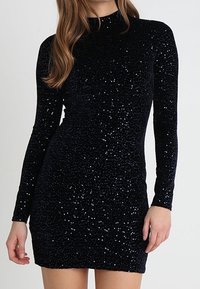 Svart paljettklänning med långa ärmar, figurnära design och hög halsringning. Tyget har en strukturerad glans från de många paljetterna.