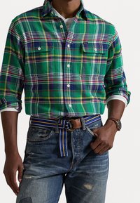 Camicia di flanella a quadri verdi con accenti viola e gialli, chiusura con bottoni, due tasche sul petto, abbinata a jeans blu consumati e una cintura a righe.