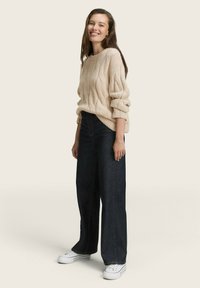 Un pull en câble crème avec des manches amples, associé à un jean large en denim sombre taille haute et des baskets blanches, le tout sur un fond neutre.