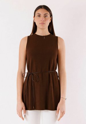 Donna in piedi, con indosso una canotta marrone con una sottile cintura annodata a fiocco in vita e pantaloni bianchi, su uno sfondo uniforme.