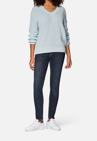 Frau trägt einen hellblauen V-Ausschnitt-Strickpullover, dunkle Skinny Jeans und weiße Sneakers und steht vor einem schlichten Hintergrund.