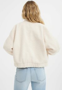 Kurtka z fakturowanego polaru w kolorze kremowym, o luźnym kroju, z opadającymi ramionami i ściągaczem u dołu. Noszona na jasnoniebieskich dżinsach.