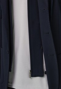 Laivastonsininen takki, jossa on sileä pinta, vetoketjukiinnitys, napit ja vinot taskut, kerrospukeutuneena valkoisen paidan päälle.