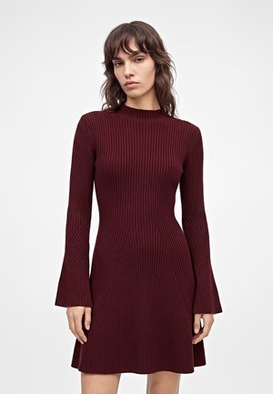 Un mannequin se tient sur un fond blanc uni, portant une robe en maille côtelée bordeaux avec de longues manches évasées et un col montant.