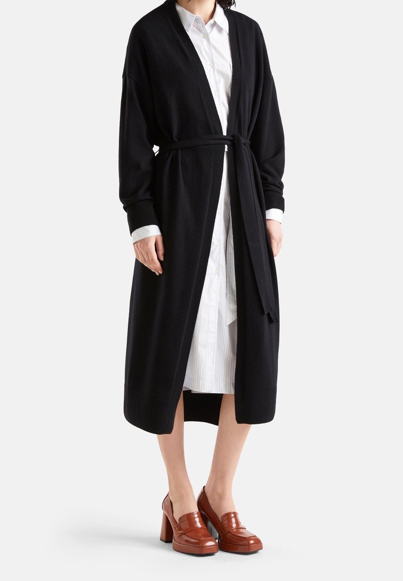 Černý dlouhý kardigan s páskem na zavazování, žebrovanými manžetami a otevřeným předním dílem, nošený přes bílou pruhovanou košili. Lesklé hnědé platformové boty.