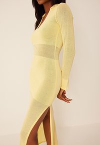 Robe jaune à manches longues en tissu maille transparent, dotée d'un décolleté en V, d'un col et d'une fente latérale. Le design présente une silhouette ajustée avec des rayures horizontales.
