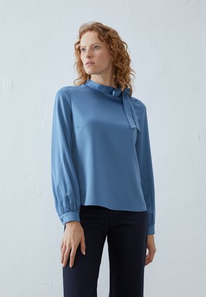 Lyseblå satengbluse med høy krage og bånddetalj, med lange, puffede ermer og en jevn tekstur. Brukt med mørke bukser.