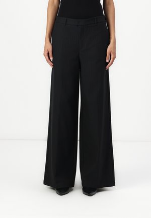 Pantalon large à fines rayures noires avec taille haute, passants pour ceinture et texture lisse, associé à des escarpins noirs pointus.