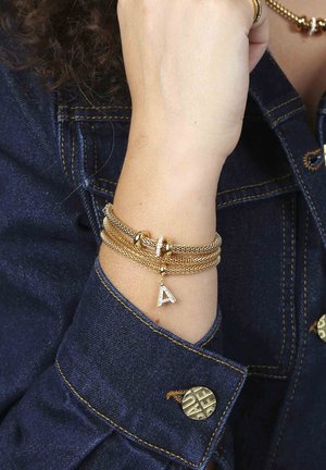 Gouden multi-strand armband met mesh ontwerp, voorzien van een kleine met diamantaccenten versierde charme in de vorm van de letter "A". Draagt om een pols tegen denim stof.