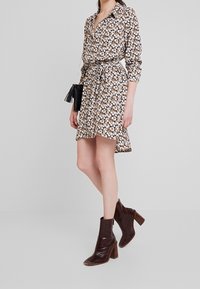 Brun blommig klänning med skjortkrage, långa ärmar, bälte i midjan och asymmetrisk nederdel. Tillsammans med bruna klackade ankelskor.