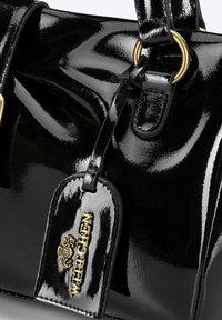 Schwarze Lackleder-Handtasche mit goldfarbenen Beschlägen, die eine glatte Textur aufweist und ein Etikett mit dem Markennamen "WITTCHEN" trägt.
