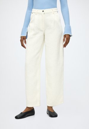 Femme portant un pantalon blanc texturé à jambes larges, un pull côtelé bleu clair avec des poignets ornés de perles et des ballerines noires, debout sur fond blanc.