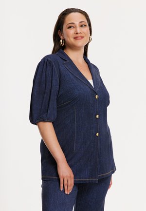 Femme portant un blazer en denim bleu foncé à manches bouffantes et un pantalon assorti, souriante avec des boucles d'oreilles créoles dorées.