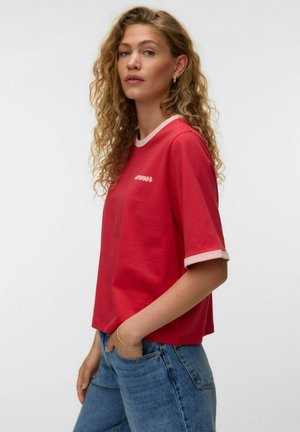 Kvinde med krøllet hår iført en rød T-shirt med hvide kanter og blå jeans, poserende med den ene hånd i lommen og neutralt udtryk.