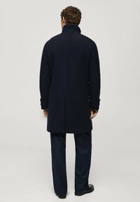 Marinblå ullkappa med hög krage och långa ärmar, med en rak passform. Tillbehör i mörka byxor och skor fulländar outfiten.