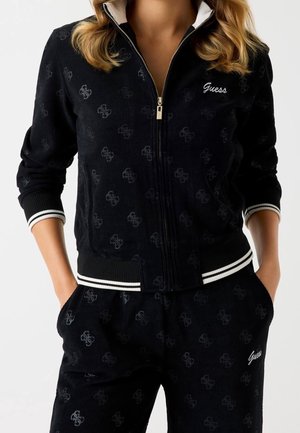 Femme portant un survêtement noir Guess avec un motif subtil de logo, une veste zippée avec un liseré blanc et un pantalon assorti, mains dans les poches.