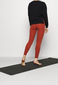 Schwarzes Oberteil mit langen Ärmeln, kombiniert mit hochtaillierten, engen kastanienbraunen Leggings. Steht auf einer schwarzen Yogamatte, mit sichtbaren barfuß Füßen.
