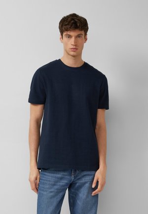 Junger Mann mit kurzen braunen Haaren, der ein einfaches dunkelblaues kurzärmliges T-Shirt und hellblaue Jeans vor grauem Hintergrund trägt.