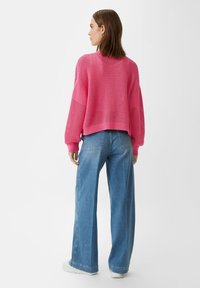 Różowy sweter dzianinowy o luźnym kroju, z teksturowanym wzorem i ściągaczami przy rękawach, zestawiony z jasnoniebieskimi spodniami typu wide-leg oraz białymi trampkami.