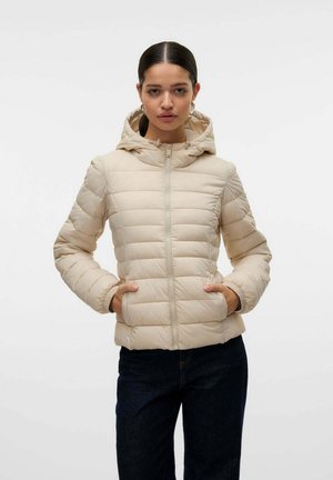 Donna con capelli scuri, indossa una giacca beige imbottita con cappuccio e pantaloni scuri, in piedi con le mani in tasca contro uno sfondo chiaro e uniforme.