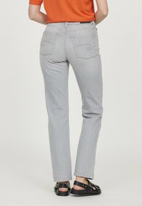 Personne portant un jean gris clair et des sandales noires, se tenant contre un fond uni, vue de dos.